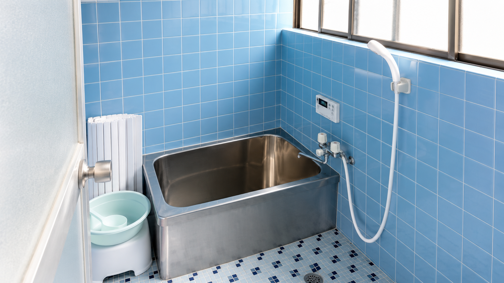Blue tiled bathroom with a rectangular stainless steel soaking tub, wall-mounted shower hose, and a blue-tiled floor and walls; small pale basin on a stool sits beside the tub.
