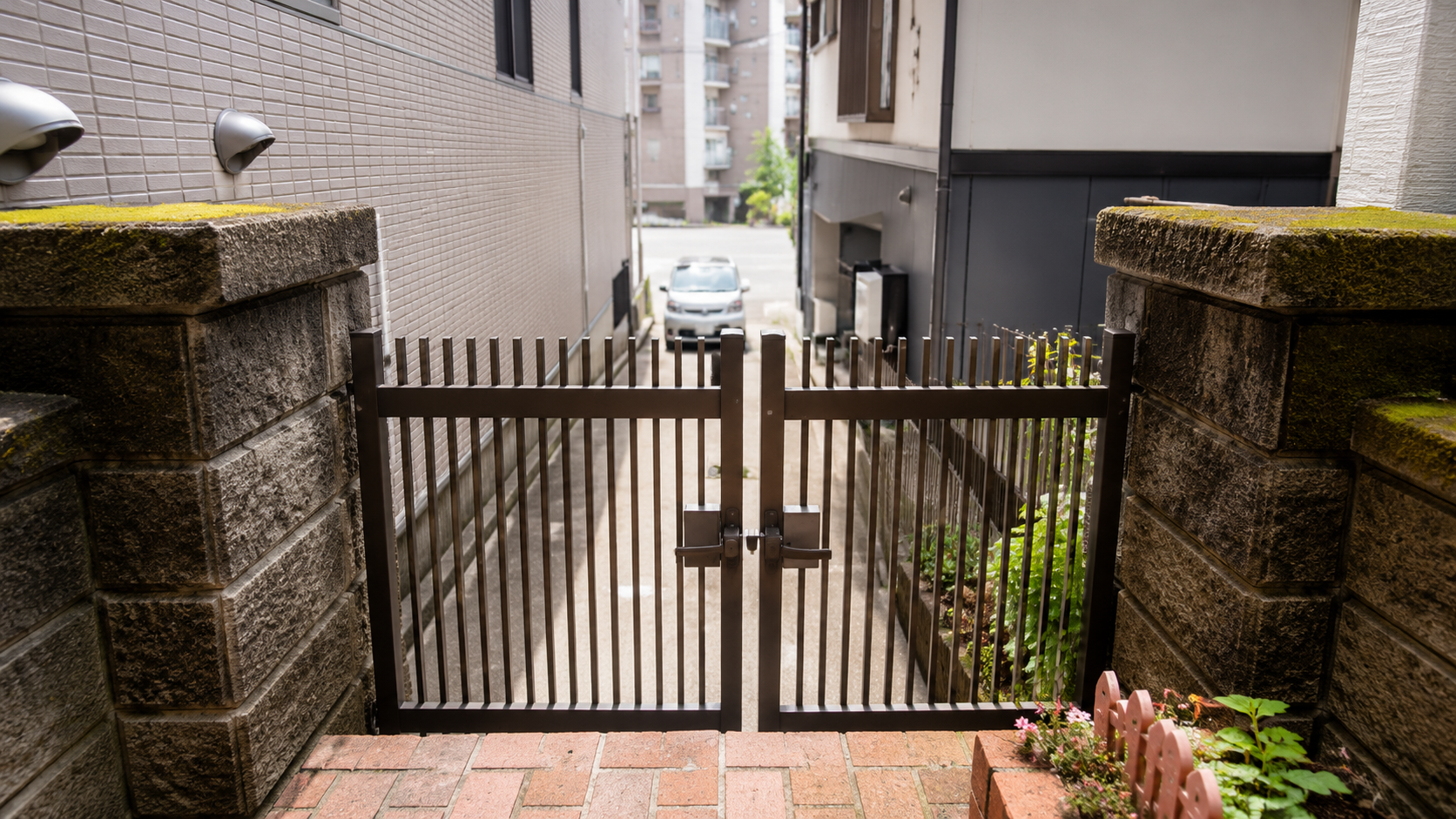 Iron vertical-bar gate at a narrow alley entrance between stone walls, leads to a street with a parked car beyond.