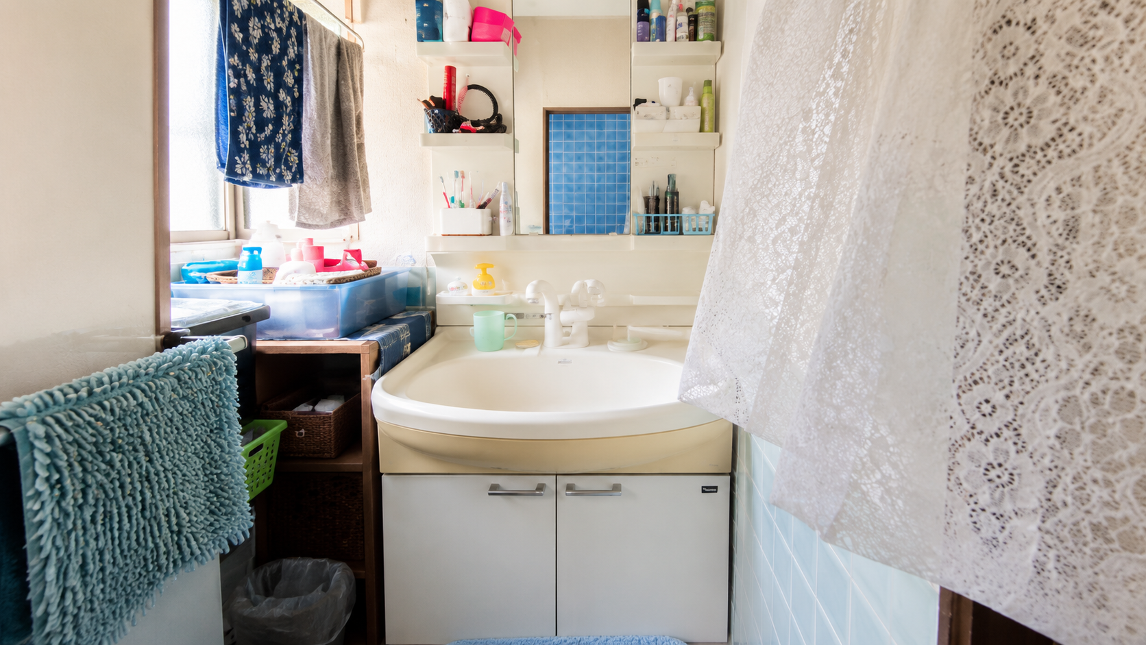 Small bathroom with a white sink, shelves, and a lace shower curtain on the right; cluttered counter with toiletries and towels nearby