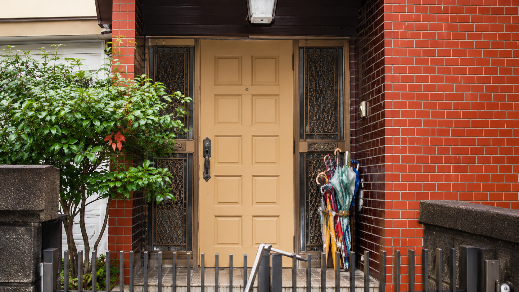 Beige paneled front door flanked by decorative iron gates, with green shrubbery on the left and a colorful umbrella stand on the right.