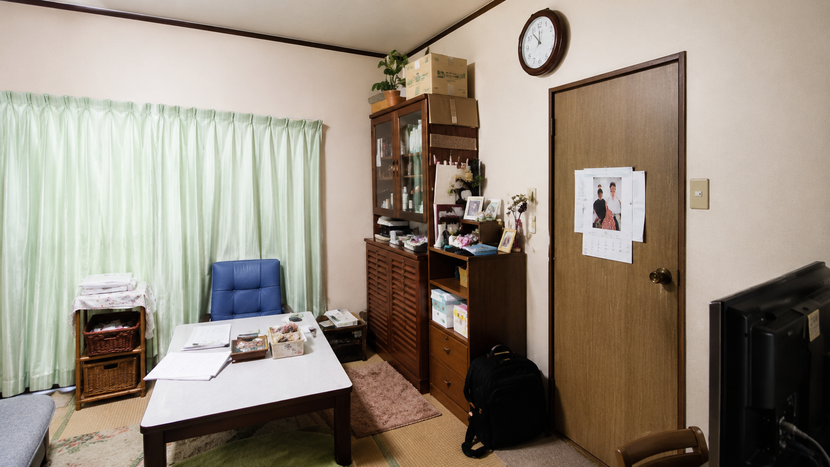 Small home office with a wooden desk, blue cushioned chair, and green curtains, plus a tall cabinet stacked with boxes on top.