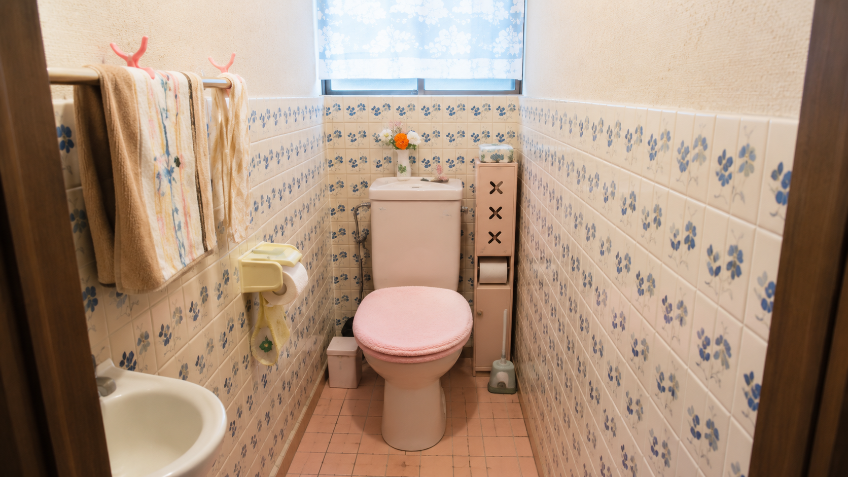 Compact vintage bathroom with a pink-seat toilet, pale pink tile floor, and blue floral wall tiles framed by a towel rack on the left.