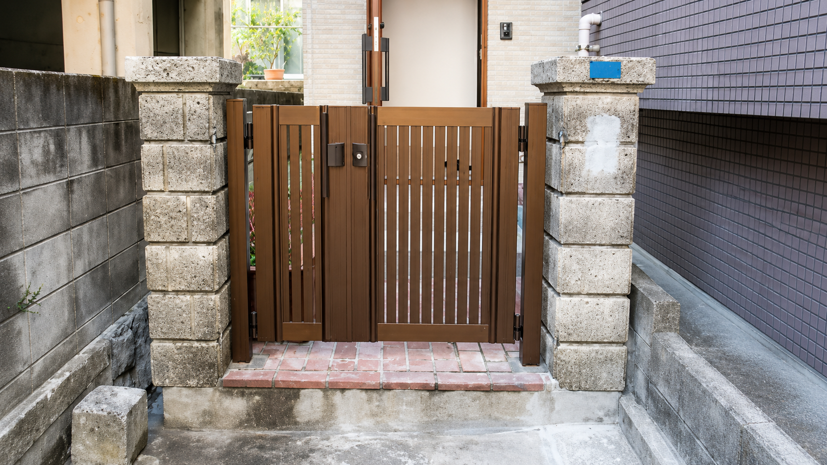 Brown wooden gate with vertical slats flanked by stone pillars at a house entrance on a brick path.
