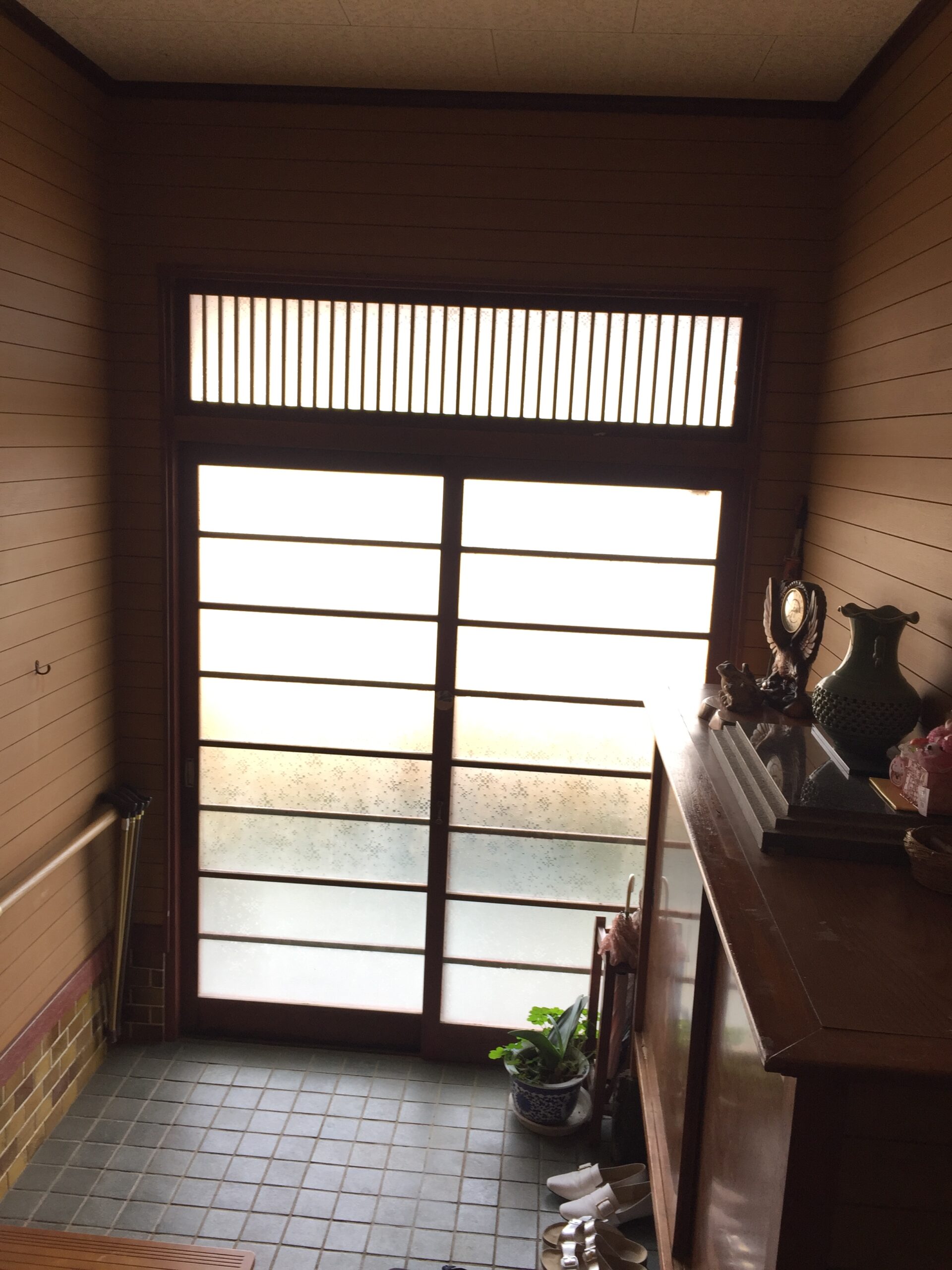 Interior entryway with a frosted glass door and a row of vertical window slats above, plus a wooden cabinet and shoes nearby on the right side of the room.