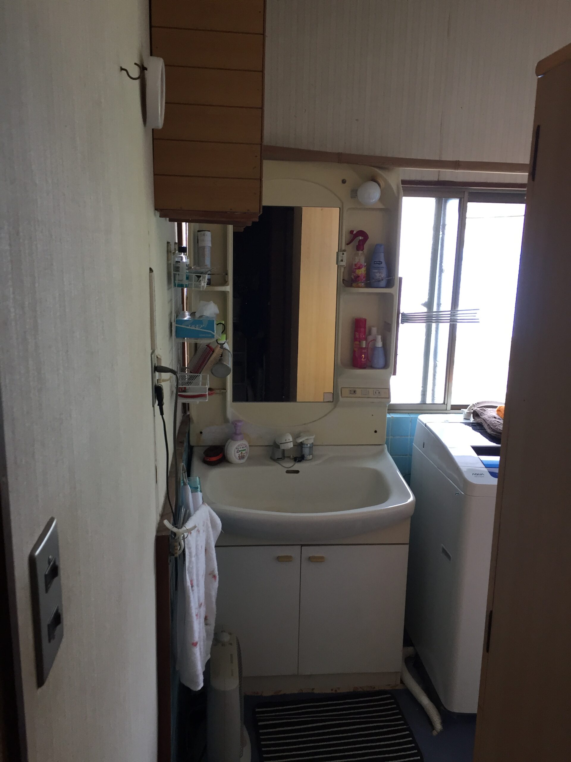 Compact bathroom with sink, mirror, and vanity; shelves hold toiletries above the sink, washing machine to the right, window near the back, and a striped rug on the floor.