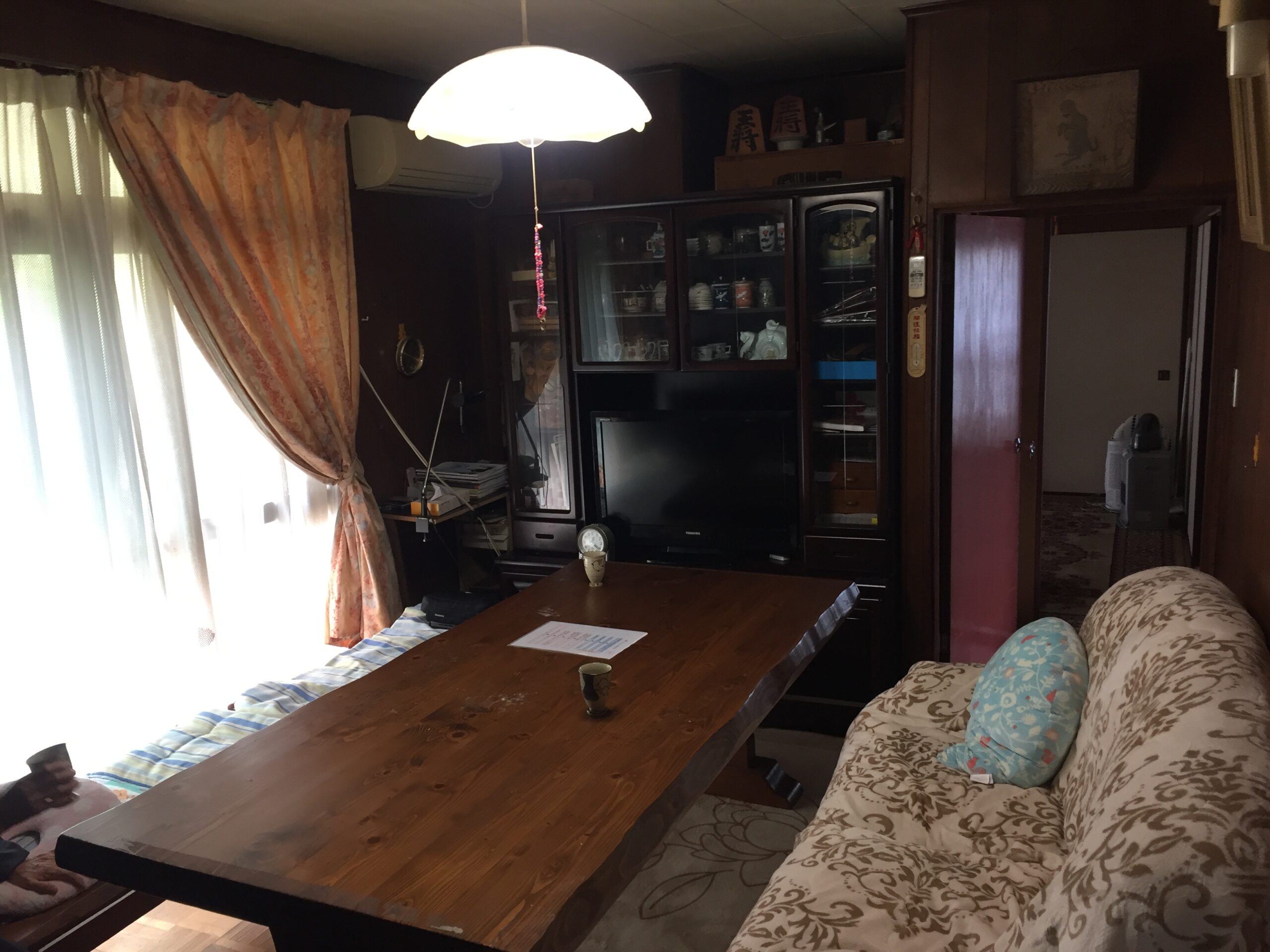 Wooden dining table in a cozy living room with floral sofa, glass-front cabinet, and warm ceiling lamp overhead.