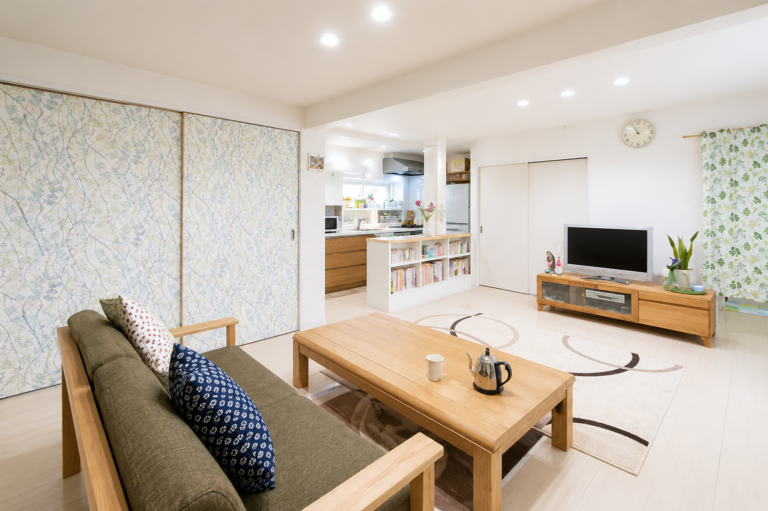 Open-plan living room and kitchen with a wooden sofa, light coffee table, and a TV on a low wooden console under a patterned floral wall; bright, cozy space.