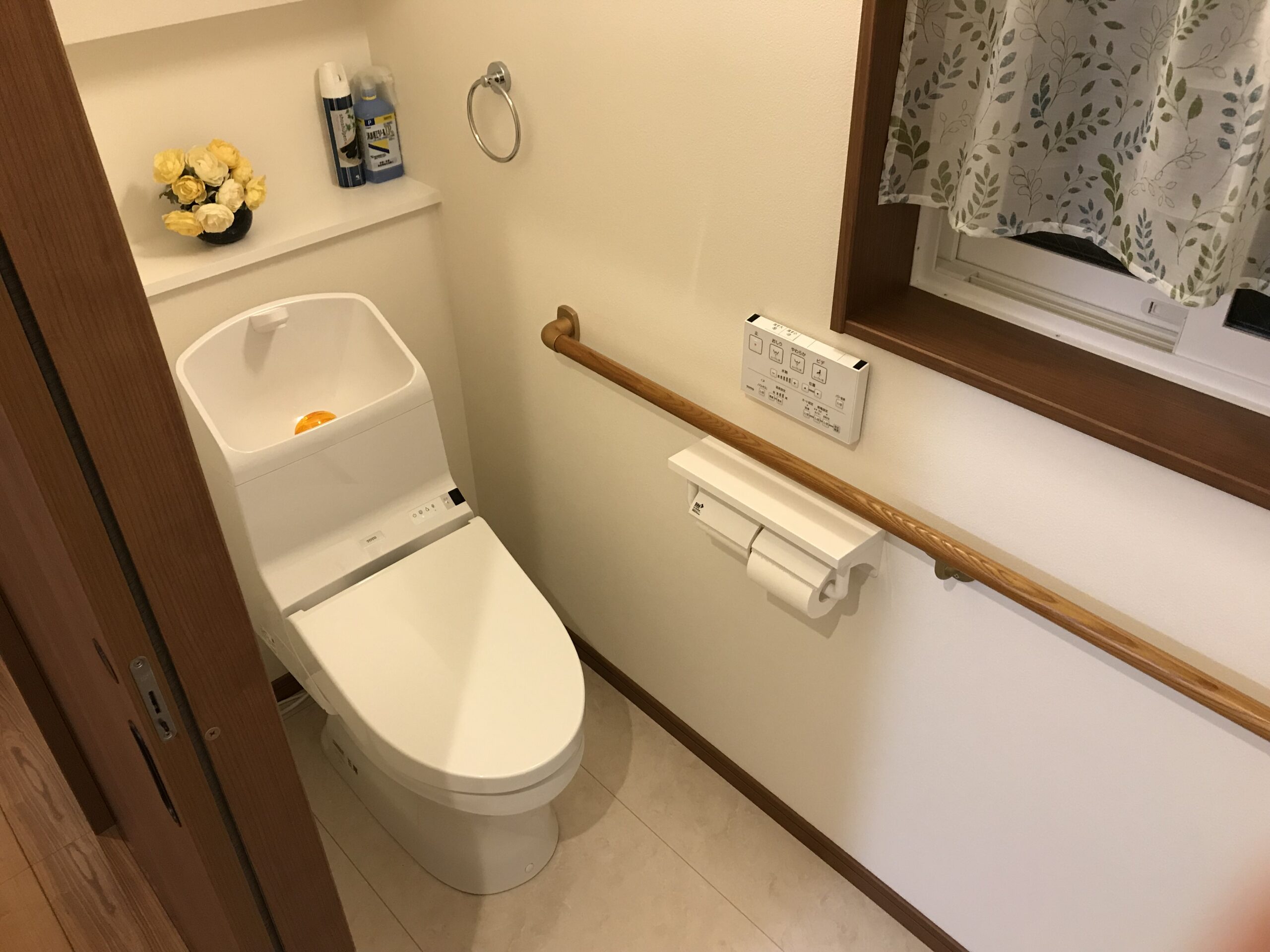 Compact bathroom with a modern bidet toilet, control panel on the wall, and a wooden handrail; shelf holds shampoo bottles and a vase of yellow and white flowers; patterned curtain at a window nearby.
