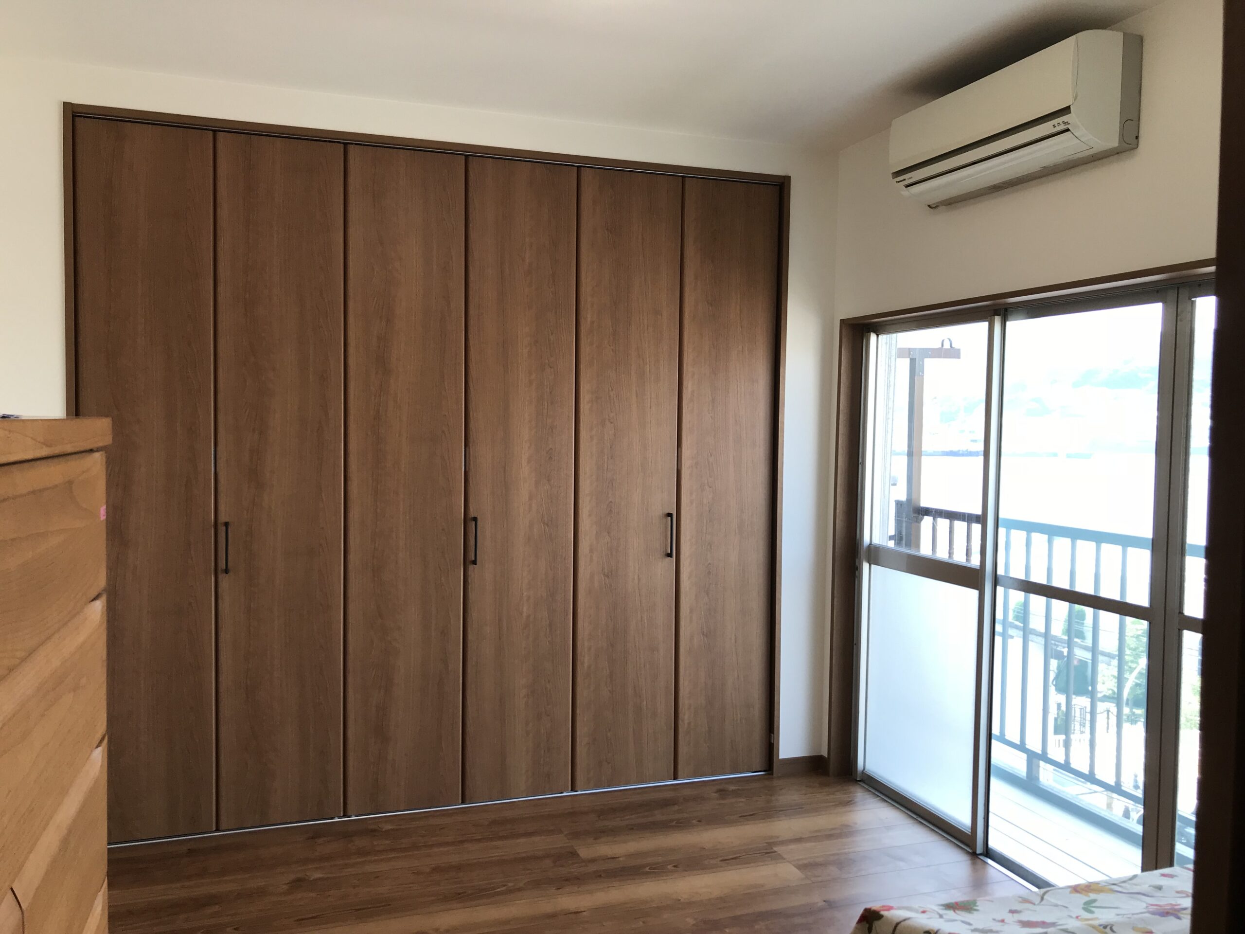 Large built-in wooden wardrobes along wall in a bright bedroom with wood flooring and a balcony window on the right; an air conditioner mounted high on the wall above.