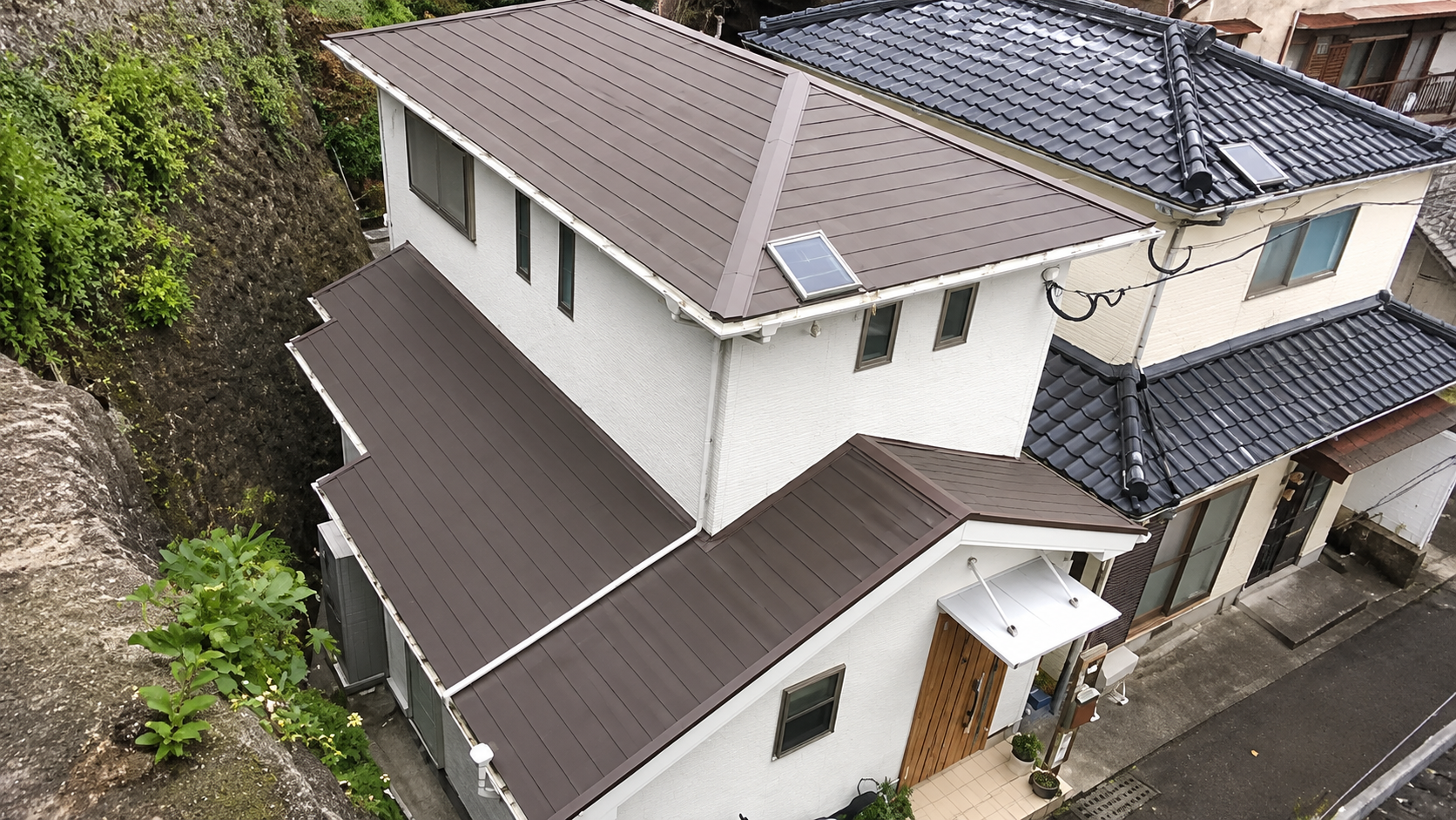 White multi-story house with brown metal roofs, a skylight, and a small entrance canopy beside a rocky hillside.