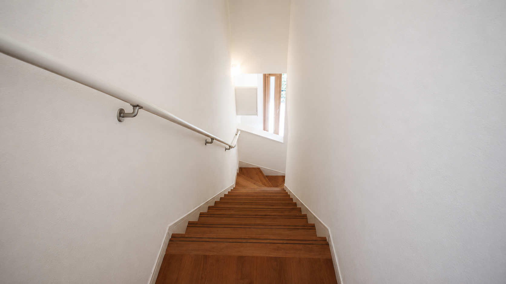 Wooden staircase descending between white walls with a metal handrail on the left, bright light at the bottom edge of the stairs.