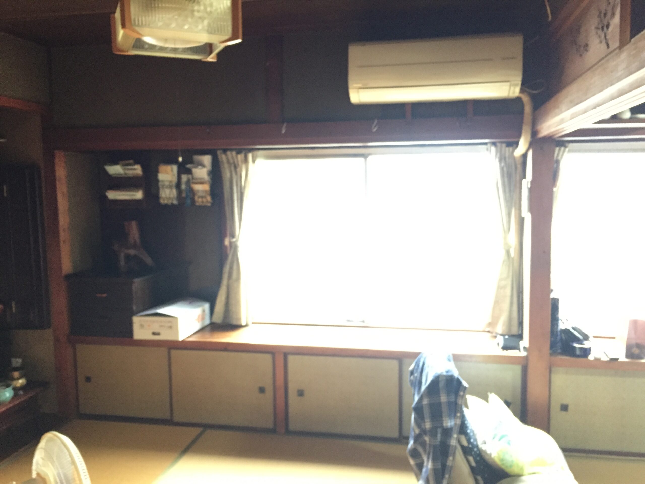 Cluttered traditional-style room with tatami floor, low cabinets, shelves, and a window behind bright light; an overhead air conditioner mounted on the wall.
