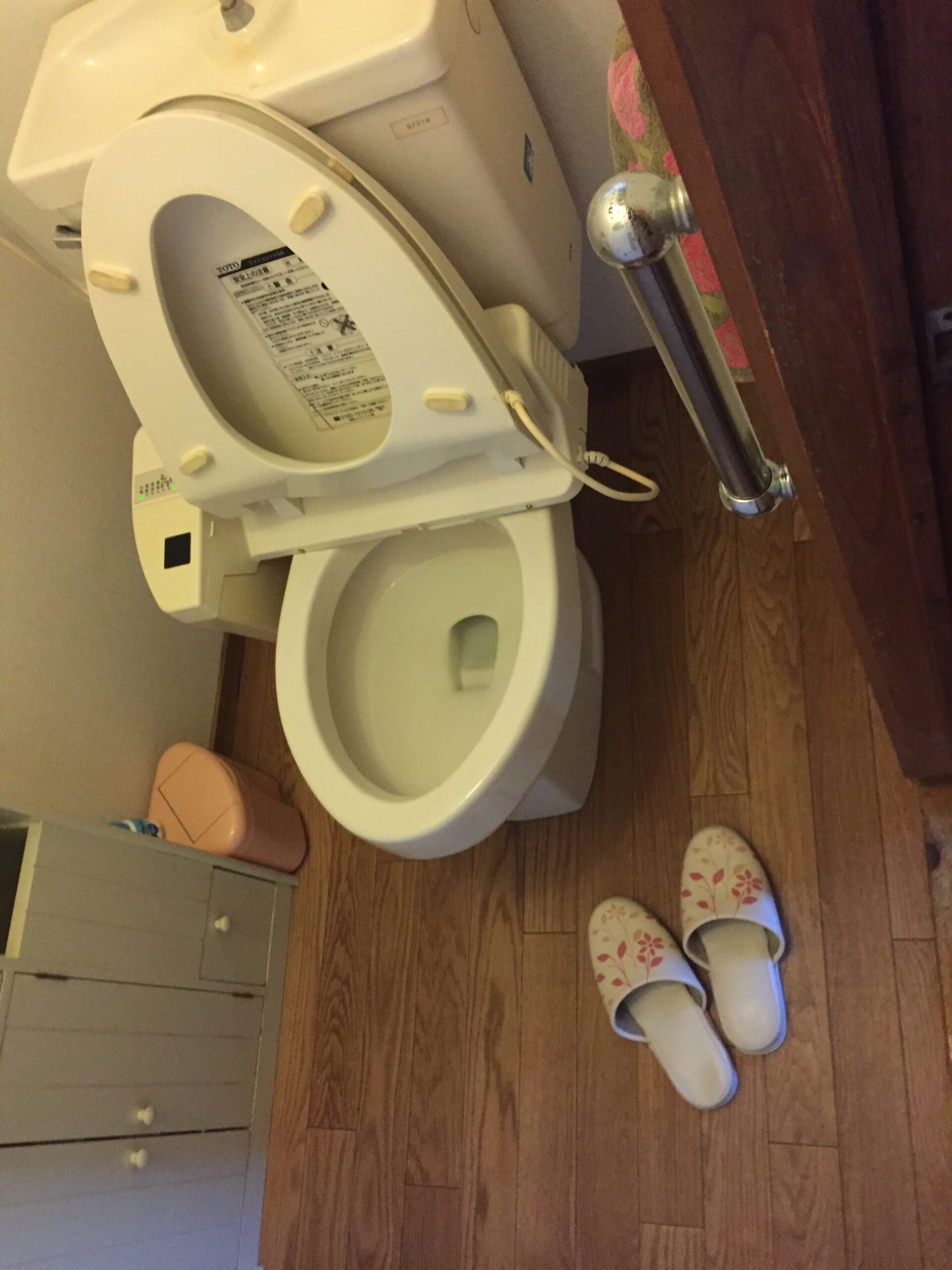 White toilet with the lid raised in a small bathroom, slippers with a floral pattern on a wooden floor nearby.