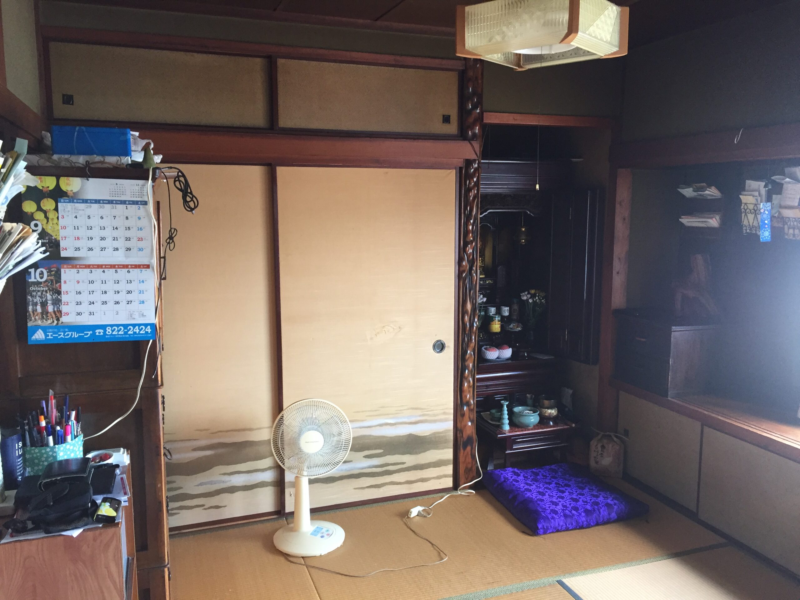 Traditional Japanese-style room with tatami mats, sliding wooden doors, and a standing pedestal fan in the center foreground; a calendar and desk items on the left wall.