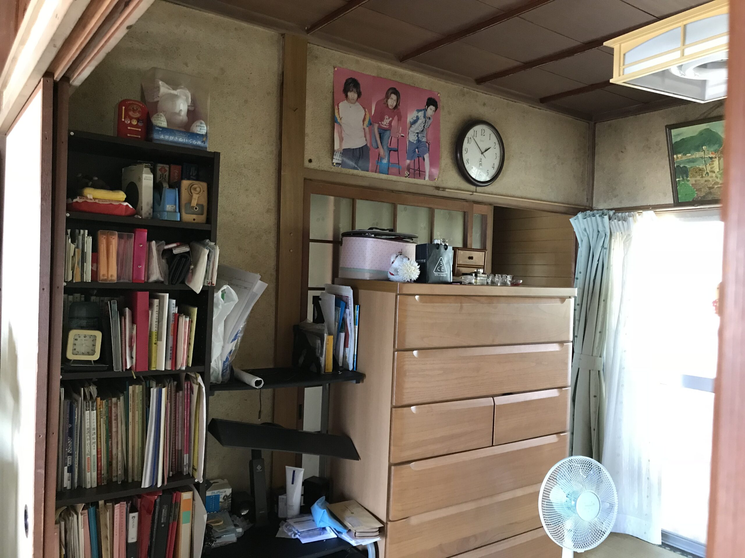 A cluttered home office or study area with tall bookshelves, stacked files, and a wooden dresser against a textured wall.
