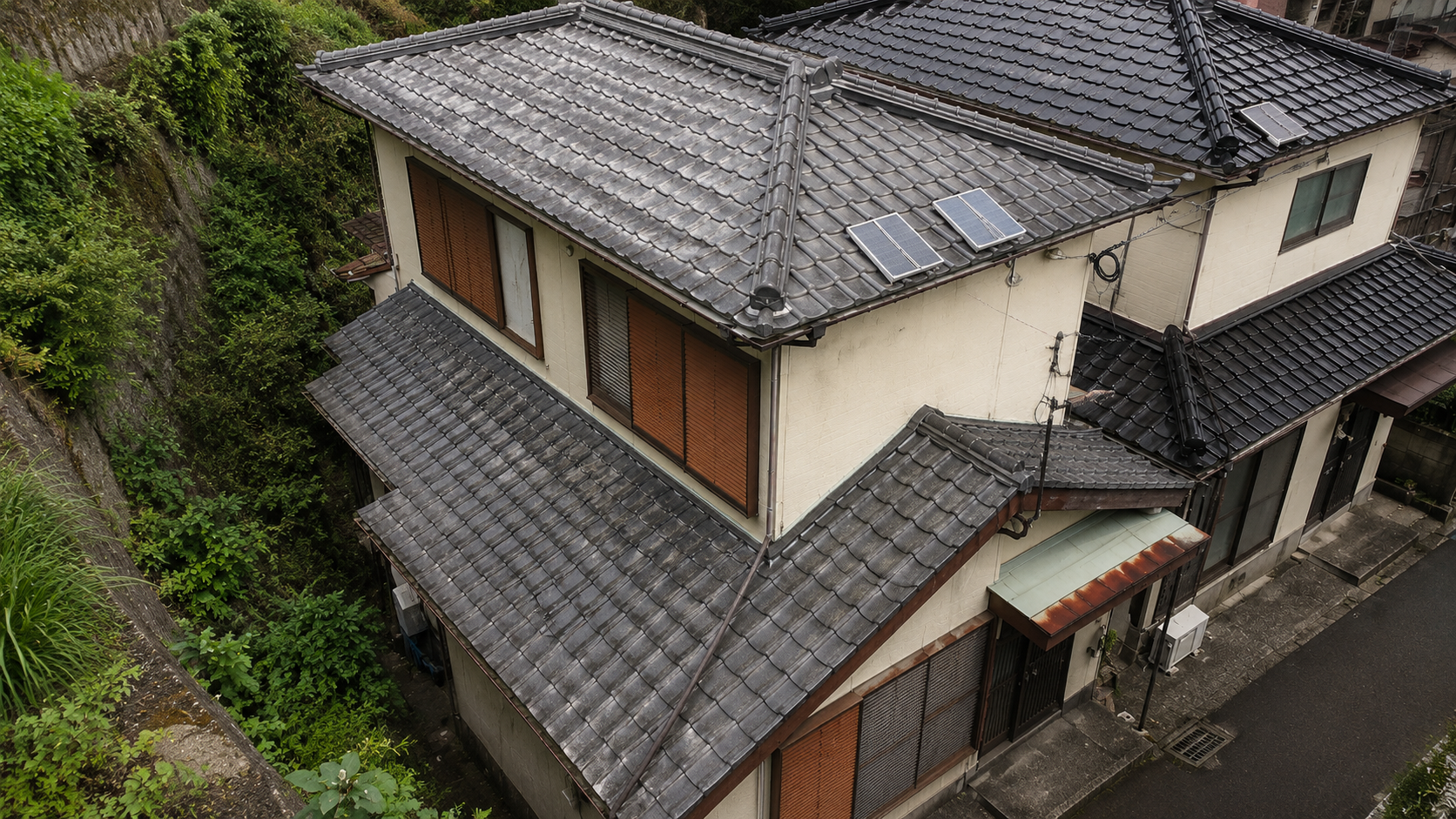 A two-story Japanese house with gray tile roof, brown shutters, beige walls, and solar panels on the upper roof, viewed from above.