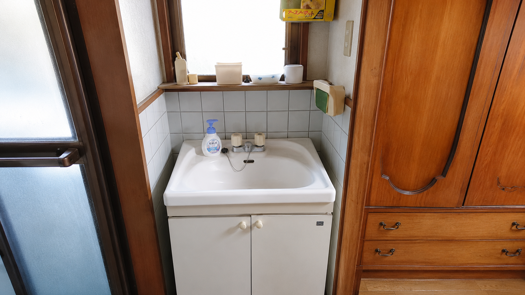 Small white bathroom sink with soap dispenser and two beige knobs, set against a tiled wall and a wooden cabinet beneath; window shelf above with containers and toothbrush items nearby.