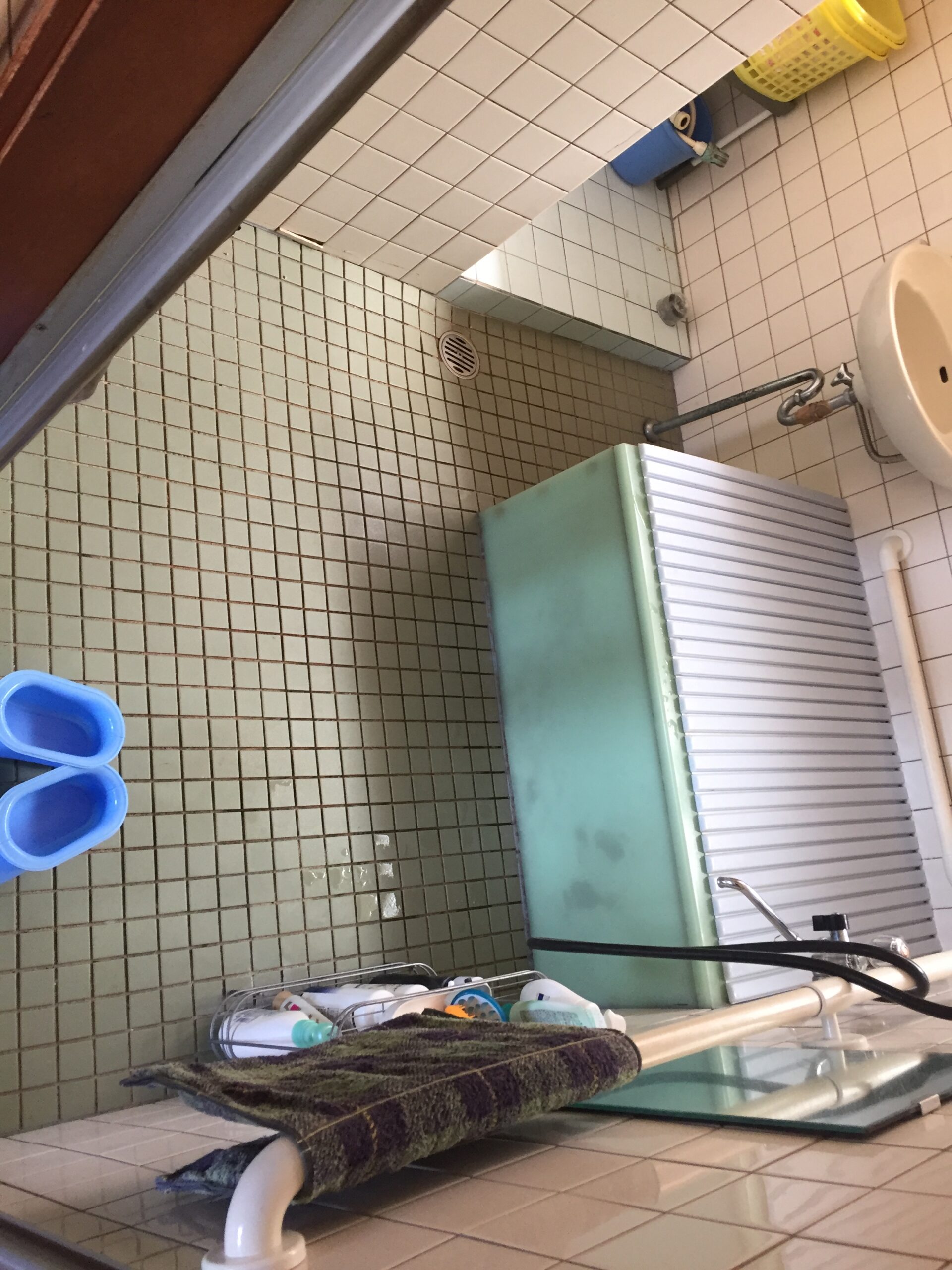 Small bathroom scene with white tiled walls, a wall-mounted sink and exposed plumbing, a mop and towel resting on a rack, and cleaning supplies in a yellow basket nearby.