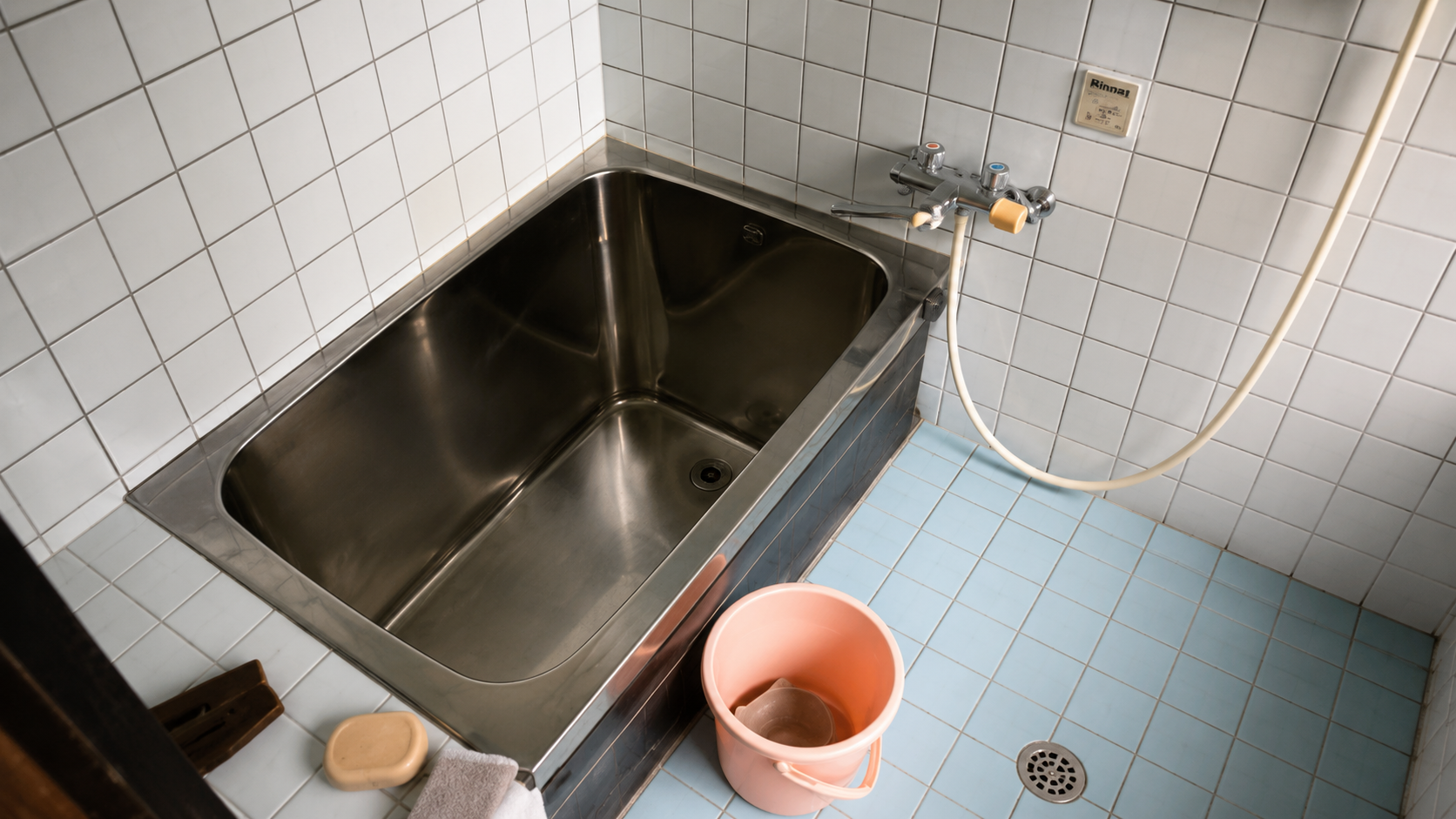 Stainless steel bathtub in a tiled bathroom with a pink bucket and cleaning supplies on the light blue floor area.