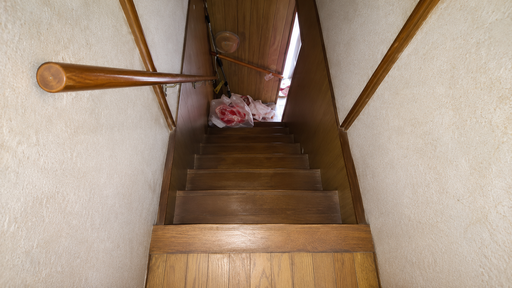 Wooden staircase viewed from above, with a handrail on the left and bags at the bottom landing.